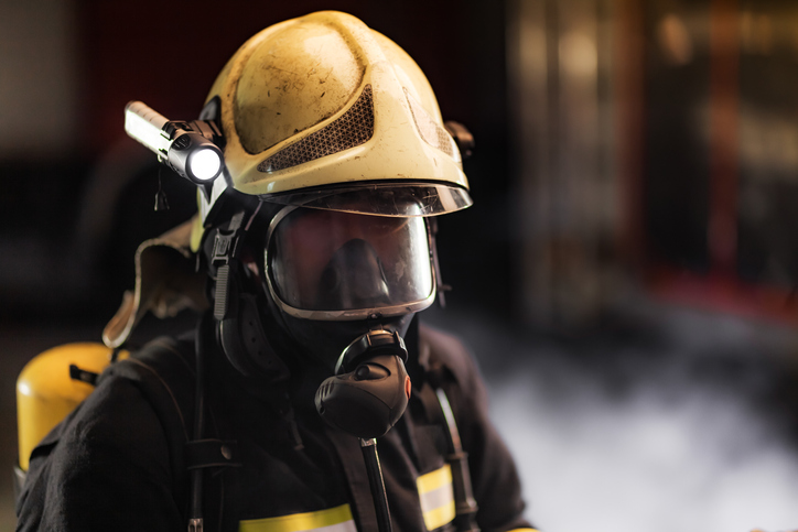 Fireman wearing full equipment. Firefighter with protective helmet oxygen mask and flashlight. Smoke and fire trucks in the background.