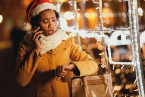 Black Woman Looking At Her Watch And Using Smartphone At Christmas Time In The City