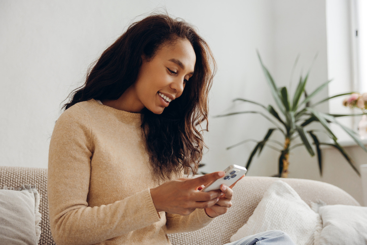 Portrait of smiling African American woman holding mobile phone using mobile app sitting on sofa